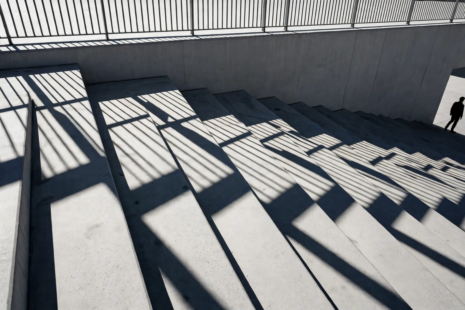 Les ombres d'une balustrade rayent de larges marches de béton en plein soleil, avec un passant en silhouette au bord du cadre